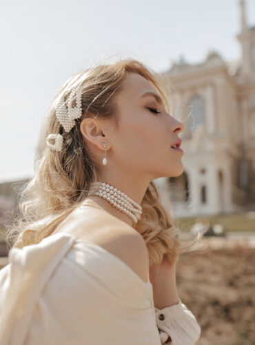 Beautiful curly lady in stylish white blouse and pearl necklace poses with closed eyes and enjoys sunny day in city center.